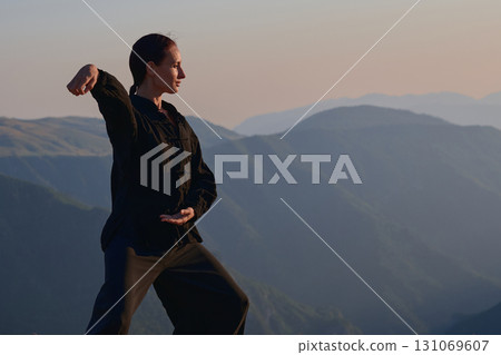 Woman practicing Tai Chi in traditional black outfit on mountain at sunrise, arms outstretched in meditation pose focusing on balance, energy, and mindfulness in nature. 131069607
