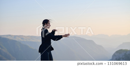 Woman practicing Tai Chi in traditional black outfit on mountain at sunrise, arms outstretched in meditation pose focusing on balance, energy, and mindfulness in nature. 131069613