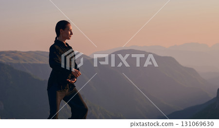 Woman practicing Tai Chi in traditional black outfit on mountain at sunrise, arms outstretched in meditation pose focusing on balance, energy, and mindfulness in nature. 131069630
