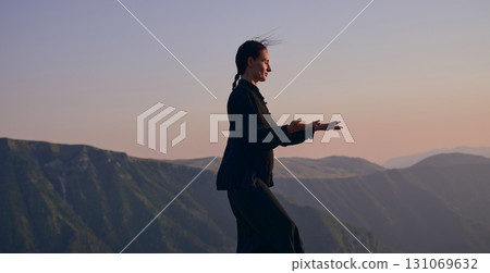 Woman practicing Tai Chi in traditional black outfit on mountain at sunrise, arms outstretched in meditation pose focusing on balance, energy, and mindfulness in nature. 131069632