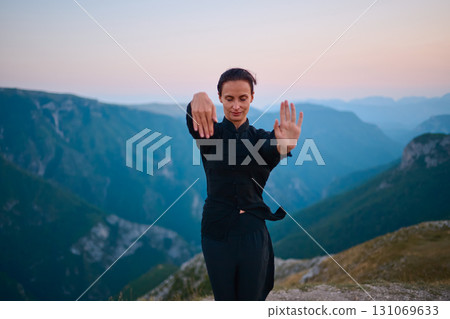 Woman practicing Tai Chi in traditional black outfit on mountain at sunrise, arms outstretched in meditation pose focusing on balance, energy, and mindfulness in nature. 131069633