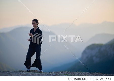 Woman practicing Tai Chi in traditional black outfit on mountain at sunrise, arms outstretched in meditation pose focusing on balance, energy, and mindfulness in nature. 131069634