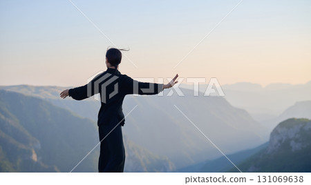 Woman practicing Tai Chi in traditional black outfit on mountain at sunrise, arms outstretched in meditation pose focusing on balance, energy, and mindfulness in nature. 131069638