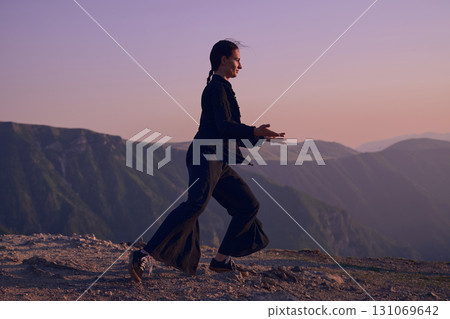 Woman practicing Tai Chi in traditional black outfit on mountain at sunrise, arms outstretched in meditation pose focusing on balance, energy, and mindfulness in nature. 131069642
