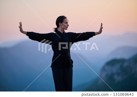 Woman practicing Tai Chi in traditional black outfit on mountain at sunrise, arms outstretched in meditation pose focusing on balance, energy, and mindfulness in nature. 131069643