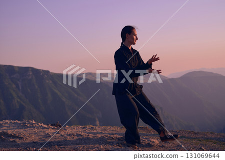 Woman practicing Tai Chi in traditional black outfit on mountain at sunrise, arms outstretched in meditation pose focusing on balance, energy, and mindfulness in nature. Woman practicing Tai Chi in traditional black outfit on mountain at sunrise, arms outstretched in meditation pose focusing on balance, energy, and mindfulness in nature. 131069644