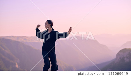 Woman practicing Tai Chi in traditional black outfit on mountain at sunrise, arms outstretched in meditation pose focusing on balance, energy, and mindfulness in nature. 131069649