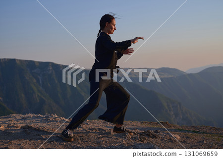 Woman practicing Tai Chi in traditional black outfit on mountain at sunrise, arms outstretched in meditation pose focusing on balance, energy, and mindfulness in nature. 131069659