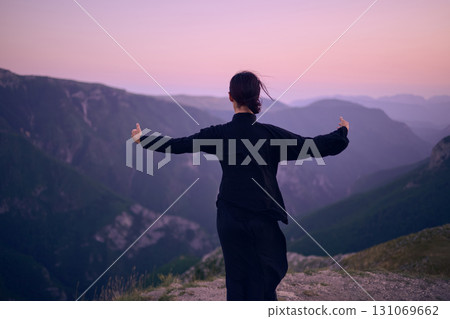 Woman practicing Tai Chi in traditional black outfit on mountain at sunrise, arms outstretched in meditation pose focusing on balance, energy, and mindfulness in nature. 131069662