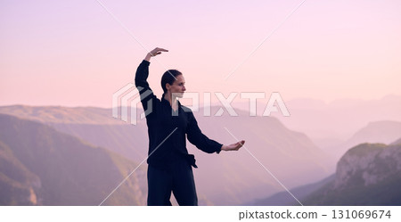 Woman practicing Tai Chi in traditional black outfit on mountain at sunrise, arms outstretched in meditation pose focusing on balance, energy, and mindfulness in nature. 131069674