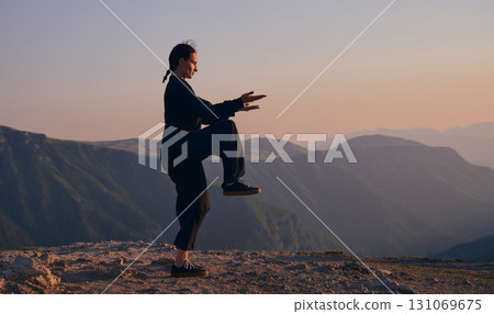 Woman practicing Tai Chi in traditional black outfit on mountain at sunrise, arms outstretched in meditation pose focusing on balance, energy, and mindfulness in nature. Woman practicing Tai Chi in traditional black outfit on mountain at sunrise, arms outstretched in meditation pose focusing on balance, energy, and mindfulness in nature. 131069675