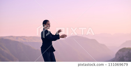 Woman practicing Tai Chi in traditional black outfit on mountain at sunrise, arms outstretched in meditation pose focusing on balance, energy, and mindfulness in nature. 131069679