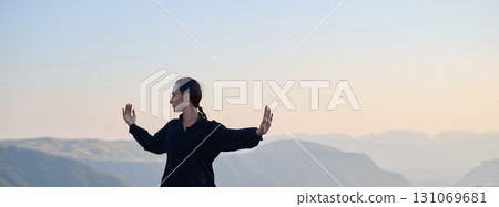 Woman practicing Tai Chi in traditional black outfit on mountain at sunrise, arms outstretched in meditation pose focusing on balance, energy, and mindfulness in nature. 131069681