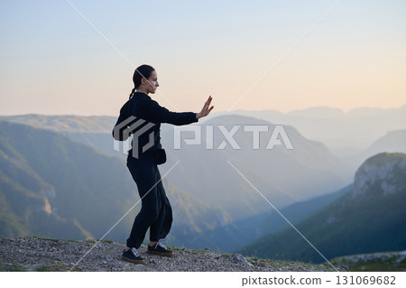 Woman practicing Tai Chi in traditional black outfit on mountain at sunrise, arms outstretched in meditation pose focusing on balance, energy, and mindfulness in nature. Woman practicing Tai Chi in traditional black outfit on mountain at sunrise, arms outstretched in meditation pose focusing on balance, energy, and mindfulness in nature. 131069682