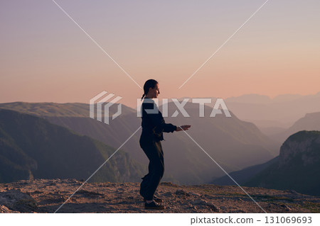 Woman practicing Tai Chi in traditional black outfit on mountain at sunrise, arms outstretched in meditation pose focusing on balance, energy, and mindfulness in nature. 131069693