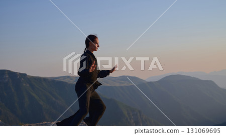 Woman practicing Tai Chi in traditional black outfit on mountain at sunrise, arms outstretched in meditation pose focusing on balance, energy, and mindfulness in nature. 131069695