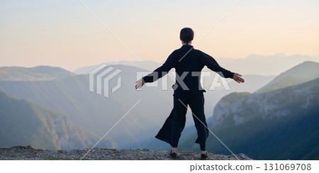 Woman practicing Tai Chi in traditional black outfit on mountain at sunrise, arms outstretched in meditation pose focusing on balance, energy, and mindfulness in nature. 131069708