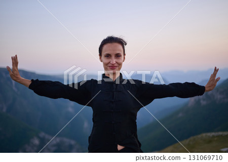 Woman practicing Tai Chi in traditional black outfit on mountain at sunrise, arms outstretched in meditation pose focusing on balance, energy, and mindfulness in nature. 131069710