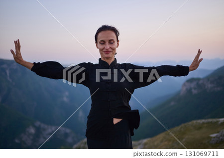 Woman practicing Tai Chi in traditional black outfit on mountain at sunrise, arms outstretched in meditation pose focusing on balance, energy, and mindfulness in nature. Woman practicing Tai Chi in traditional black outfit on mountain at sunrise, arms outstretched in meditation pose focusing on balance, energy, and mindfulness in nature. 131069711