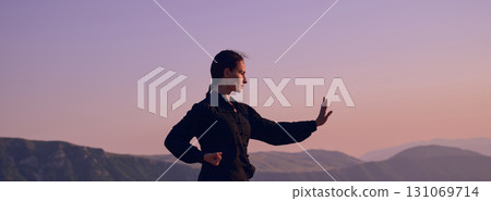 Woman practicing Tai Chi in traditional black outfit on mountain at sunrise, arms outstretched in meditation pose focusing on balance, energy, and mindfulness in nature. 131069714