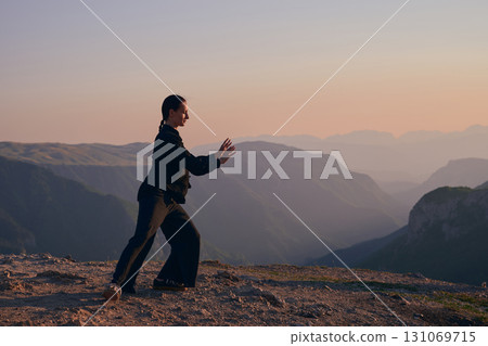Woman practicing Tai Chi in traditional black outfit on mountain at sunrise, arms outstretched in meditation pose focusing on balance, energy, and mindfulness in nature. 131069715