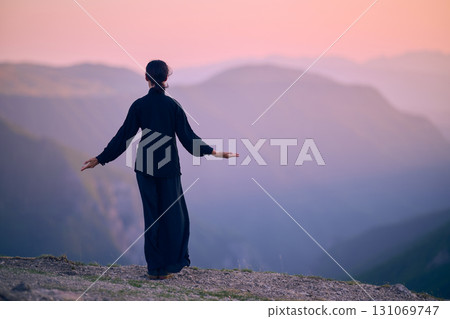 Woman practicing Tai Chi in traditional black outfit on mountain at sunrise, arms outstretched in meditation pose focusing on balance, energy, and mindfulness in nature. 131069747