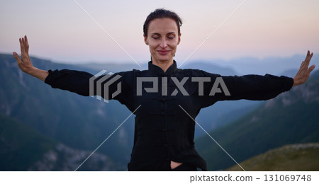 Woman practicing Tai Chi in traditional black outfit on mountain at sunrise, arms outstretched in meditation pose focusing on balance, energy, and mindfulness in nature. 131069748