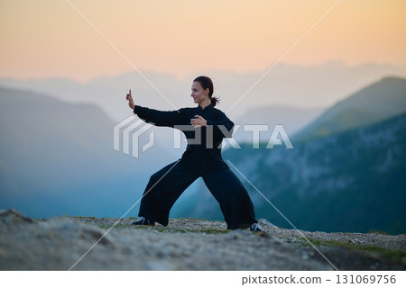 Woman practicing Tai Chi in traditional black outfit on mountain at sunrise, arms outstretched in meditation pose focusing on balance, energy, and mindfulness in nature. 131069756