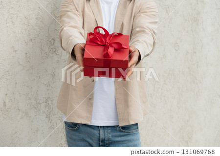 Man giving red gift box tied with ribbon for Birthday or Valentine's Day and stand against concrete beige wall. Close up Man giving red gift box tied with ribbon for Birthday or Valentine's Day and stand against concrete beige wall. Close up 131069768