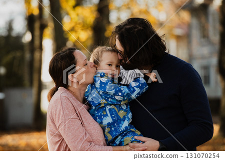 On a sunny autumn day in the park, a mother and father lovingly embrace their young son, sharing moments of laughter and joy as they play together 131070254