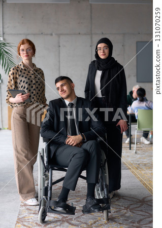 A diverse business team poses confidently, including a smiling director seated in a wheelchair, flanked by two professional female colleagues, one wearing a hijab and the other holding a tablet, in a 131070259