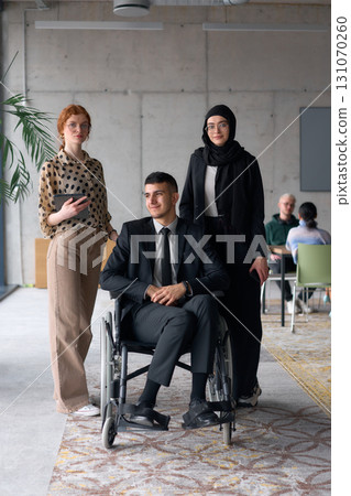A diverse business team poses confidently, including a smiling director seated in a wheelchair, flanked by two professional female colleagues, one wearing a hijab and the other holding a tablet, in a 131070260