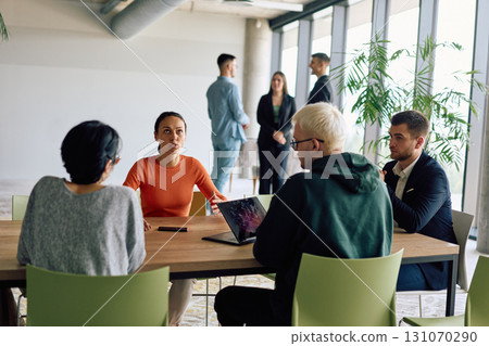 A diverse team of business professionals engaged in a discussion around a conference table in an office, while their colleagues collaborate in the background A diverse team of business professionals engaged in a discussion around a conference table in an office, while their colleagues collaborate in the background 131070290