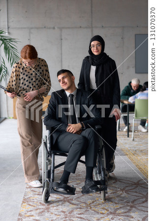 A diverse business team poses confidently, including a smiling director seated in a wheelchair, flanked by two professional female colleagues, one wearing a hijab and the other holding a tablet, in a 131070370