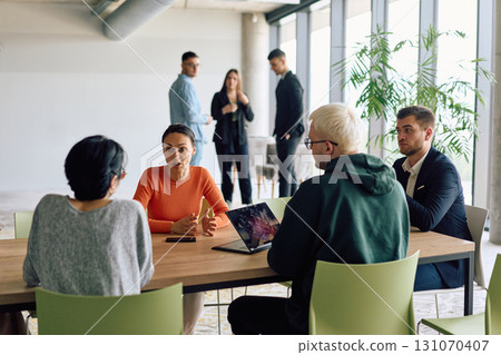 A diverse team of business professionals engaged in a discussion around a conference table in an office, while their colleagues collaborate in the background A diverse team of business professionals engaged in a discussion around a conference table in an office, while their colleagues collaborate in the background 131070407