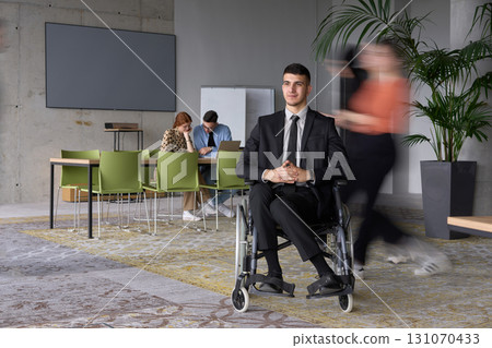 A businessman in a wheelchair navigating through a busy office, surrounded by his colleagues who are actively engaged in their work and collaboration 131070433