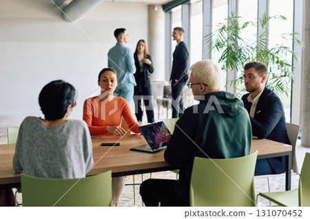 A diverse team of business professionals engaged in a discussion around a conference table in an office, while their colleagues collaborate in the background A diverse team of business professionals engaged in a discussion around a conference table in an office, while their colleagues collaborate in the background 131070452