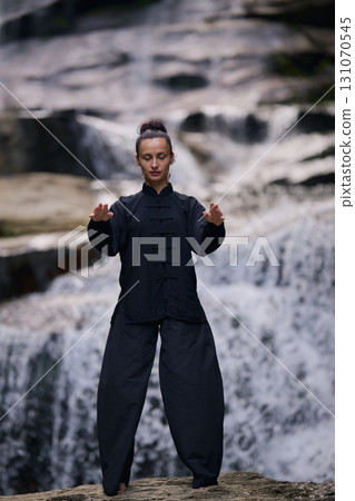 Woman practicing Tai Chi in nature, wearing a traditional black martial arts uniform, standing gracefully near a waterfall, focused on meditation and balance. Healthy lifestyle in asian culture 131070545