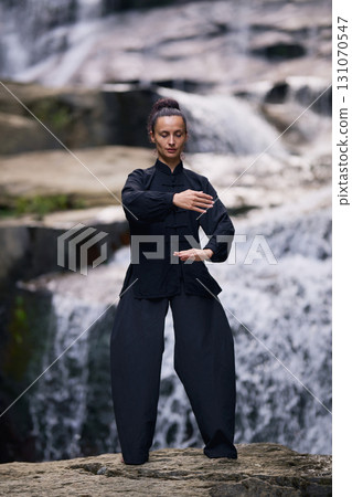 Woman practicing Tai Chi in nature, wearing a traditional black martial arts uniform, standing gracefully near a waterfall, focused on meditation and balance. Healthy lifestyle in asian culture 131070547