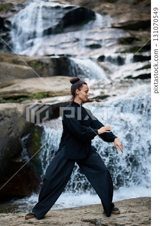 Beautiful young woman performing slow tai chi movements near a waterfall and river at sunset capturing harmony balance and fitness in nature. 131070549