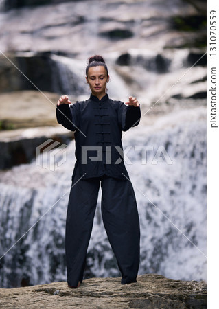 Woman practicing Tai Chi in nature, wearing a traditional black martial arts uniform, standing gracefully near a waterfall, focused on meditation and balance. Healthy lifestyle in asian culture 131070559
