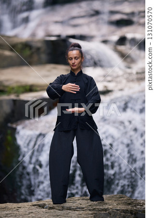 Woman practicing Tai Chi in nature, wearing a traditional black martial arts uniform, standing gracefully near a waterfall, focused on meditation and balance. Healthy lifestyle in asian culture Woman practicing Tai Chi in nature, wearing a traditional black martial arts uniform, standing gracefully near a waterfall, focused on meditation and balance. Healthy lifestyle in asian culture 131070570