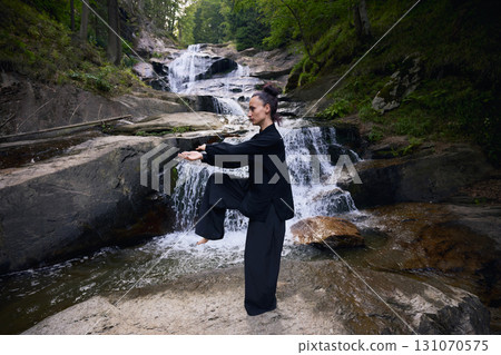 Woman practicing Tai Chi in nature, wearing a traditional black martial arts uniform, standing gracefully near a waterfall, focused on meditation and balance. Healthy lifestyle in asian culture 131070575
