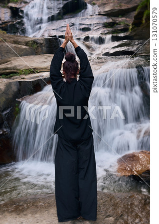 Woman practicing Tai Chi in nature, wearing a traditional black martial arts uniform, standing gracefully near a waterfall, focused on meditation and balance. Healthy lifestyle in asian culture Woman practicing Tai Chi in nature, wearing a traditional black martial arts uniform, standing gracefully near a waterfall, focused on meditation and balance. Healthy lifestyle in asian culture 131070579
