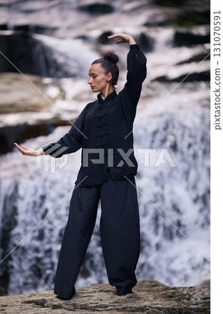 Woman practicing Tai Chi in nature, wearing a traditional black martial arts uniform, standing gracefully near a waterfall, focused on meditation and balance. Healthy lifestyle in asian culture Woman practicing Tai Chi in nature, wearing a traditional black martial arts uniform, standing gracefully near a waterfall, focused on meditation and balance. Healthy lifestyle in asian culture 131070591