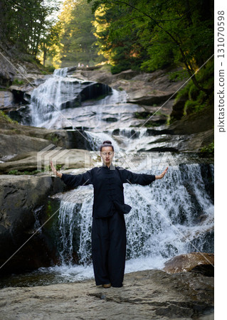 Beautiful young woman performing slow tai chi movements near a waterfall and river at sunset capturing harmony balance and fitness in nature. Beautiful young woman performing slow tai chi movements near a waterfall and river at sunset capturing harmony balance and fitness in nature. 131070598