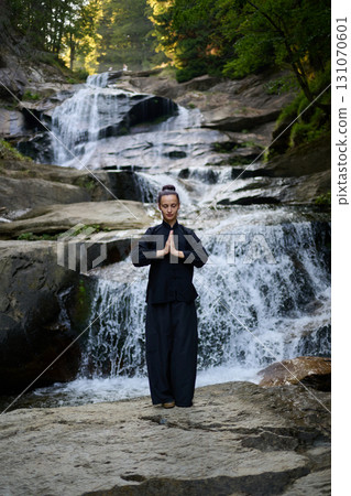 Beautiful young woman performing slow tai chi movements near a waterfall and river at sunset capturing harmony balance and fitness in nature. 131070601