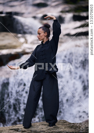 Woman practicing Tai Chi in nature, wearing a traditional black martial arts uniform, standing gracefully near a waterfall, focused on meditation and balance. Healthy lifestyle in asian culture Woman practicing Tai Chi in nature, wearing a traditional black martial arts uniform, standing gracefully near a waterfall, focused on meditation and balance. Healthy lifestyle in asian culture 131070603