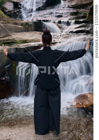 Woman practicing Tai Chi in nature, wearing a traditional black martial arts uniform, standing gracefully near a waterfall, focused on meditation and balance. Healthy lifestyle in asian culture 131070606
