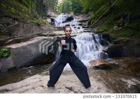 Woman practicing Tai Chi in nature, wearing a traditional black martial arts uniform, standing gracefully near a waterfall, focused on meditation and balance. Healthy lifestyle in asian culture 131070613